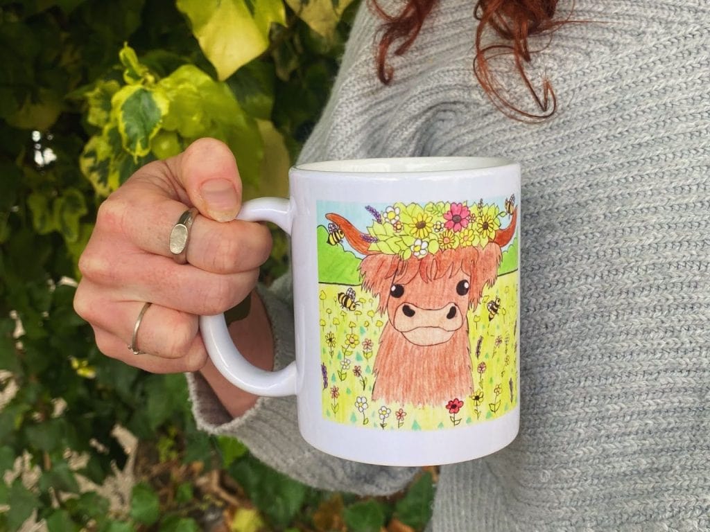 A person holding a mug decorated with a colorful drawing of a Highland cow wearing a flower crown, surrounded by bees and flowers.